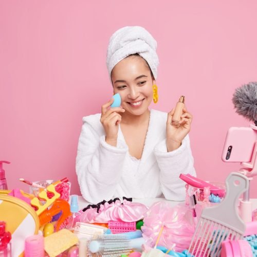 woman-with-happy-expression-applies-foundation-face-uses-socail-media-marketing-records-video-her-beauty-blog-poses-smartphone-camera-isolated-pink-wall-online-translation-scaled.jpg